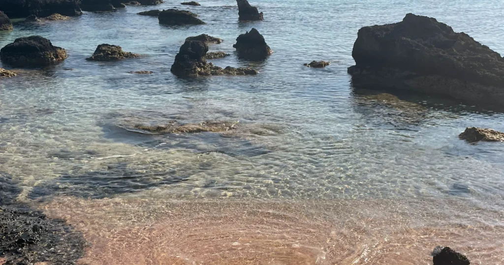 Elafonisi Beach image of edge of water with pink sand with large rocks in the background