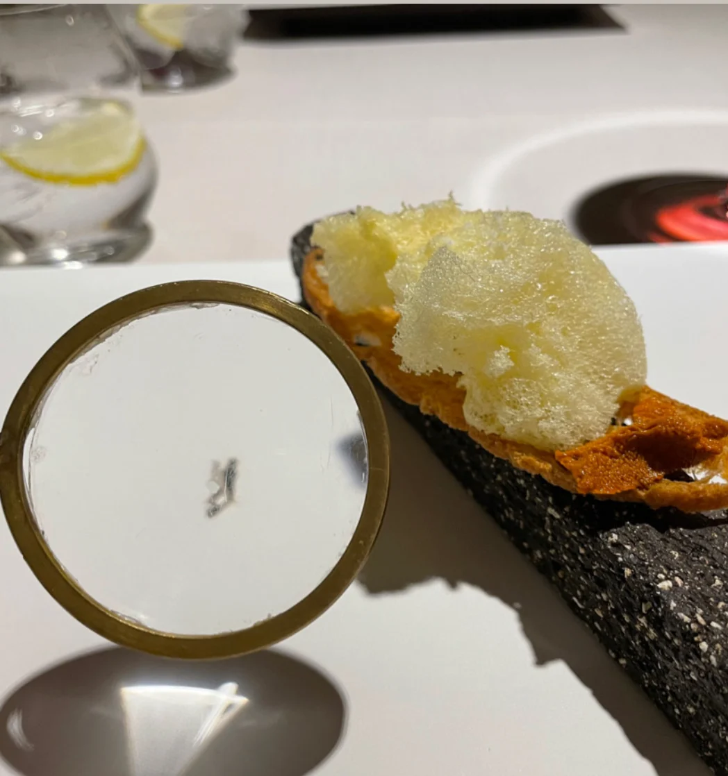 Disfrutar 32 Course Tasting Menu image of butter on a cracker on a black stone base and a magnifying glass focused on a small cutout of a man