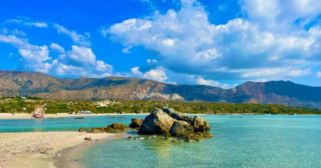 Elafonisi Beach with clear turquoise waters, sand and a pile of rocks in the foreground, blue skies with clouds and mountains in the background
