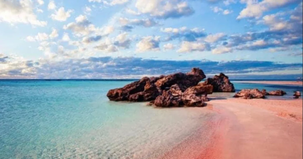 Elafonisi Beach image with pink sand to the right and clear blue water to the left and some pinkish brown rocks in the middle with blue skies and puffy white clouds