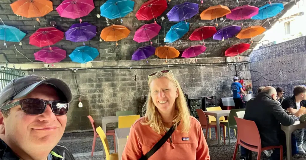 Two traveling after one seated at an outdoor market under a bridge with colorful umbrellas hanging from the ceiling.