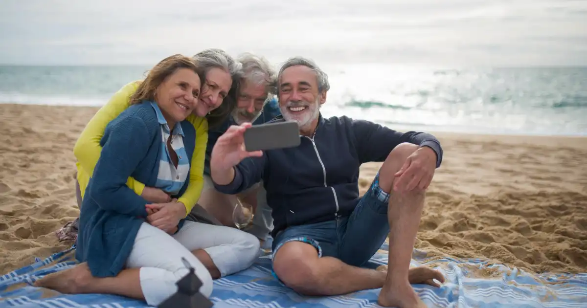 four seniors sitting on a blue blanket at the beach taking a selfie and using Travel Technology